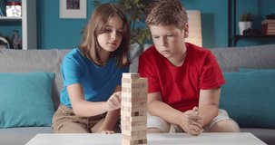 Cheerful brother and sister in casual clothing picking one by one wooden bricks from tower while sitting on couch. Smiling children playing with wooden toys at home.
