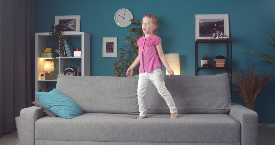 Pretty young child with ponytail dancing and singing on grey couch at living room. Happy girl in casual clothing having full of energy while staying at home.
