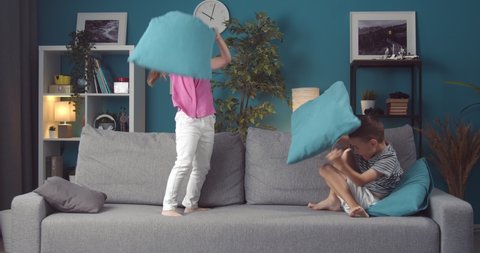 Smiling little kids in casual outfit fighting with blue pillows on grey couch. Happy sister and brother taking fun at home. Concept of entertainment.