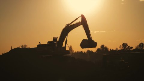 Silhouette of an excavator that loads sand into a truck at sunset. Concept construction and heavy industry, machine will be used in heavy industry business. Slow motion footage.