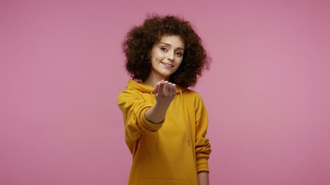 Come here, follow me! Beautiful young woman afro hairstyle in hoodie making beckoning gesture with one finger, inviting to approach, looking playful flirting. indoor   isolated on pink background