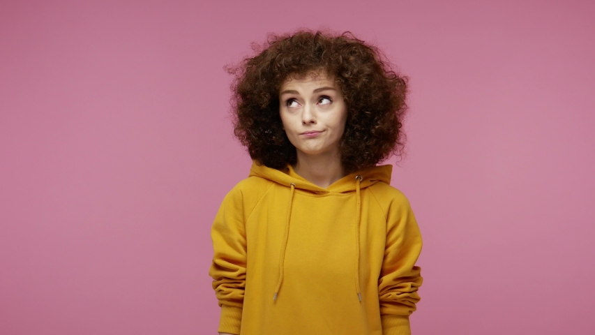 Thoughtful bewildered young woman afro hairstyle in hoodie looking big eyes amazed confused expression, having doubt hesitation, can't make decision, difficult choice.  isolated on pink background Royalty-Free Stock Footage #1058259346