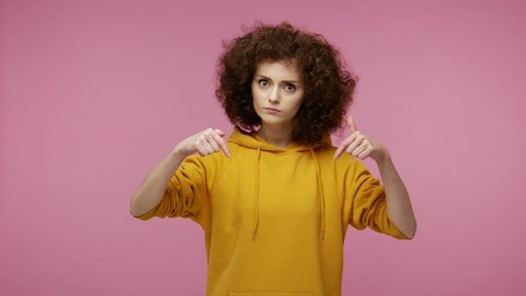 Here and right now! Angry strict young woman afro hairstyle in hoodie pointing down and looking bossy, demanding to do immediately, control in relationships. indoor   isolated on pink background