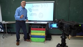 Male teacher using an interactive whiteboard in an empty classroom teaching to a video camera.