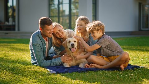 Smiling Portrait of Beautiful Family of Four Having Picnic on the Lawn, Posing with Happy Golden Retriever Dog. Idyllic Family Have Fun with Loyal Pedigree Dog Outdoors in Summer House Backyard