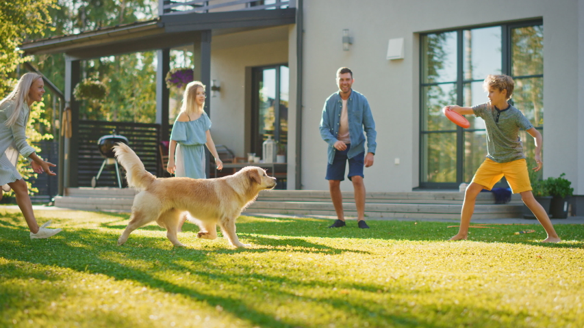 Smiling Beautiful Family of Four Play Fetch Frisbee with Happy Golden Retriever Dog on the Backyard Lawn. Idyllic Family Has Fun with Loyal Pedigree Dog Outdoors in Summer House Backyard. Slow Motion Royalty-Free Stock Footage #1058261986