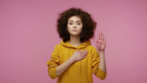 Honest responsible young woman afro hairstyle in hoodie raising hand, touching chest and taking vow, saying promise, looking at camera trustworthy reliable. indoor  isolated on pink background