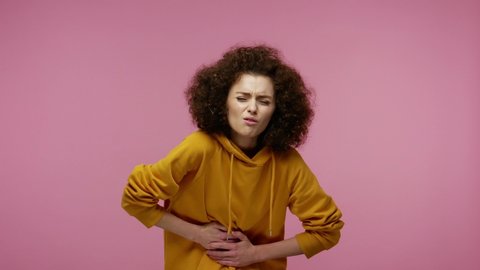 Acute abdominal cramps. Sick young woman afro hairstyle in hoodie suffering stomach ache, feeling heartburn, discomfort from constipation, gastrointestinal problem.   isolated on pink background