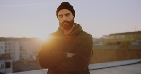 Portrait attractive adult bearded man on rooftop at sunset wearing jacket and beanie hat looking confident in urban city background.Carefree holiday and happy active lifestyle.