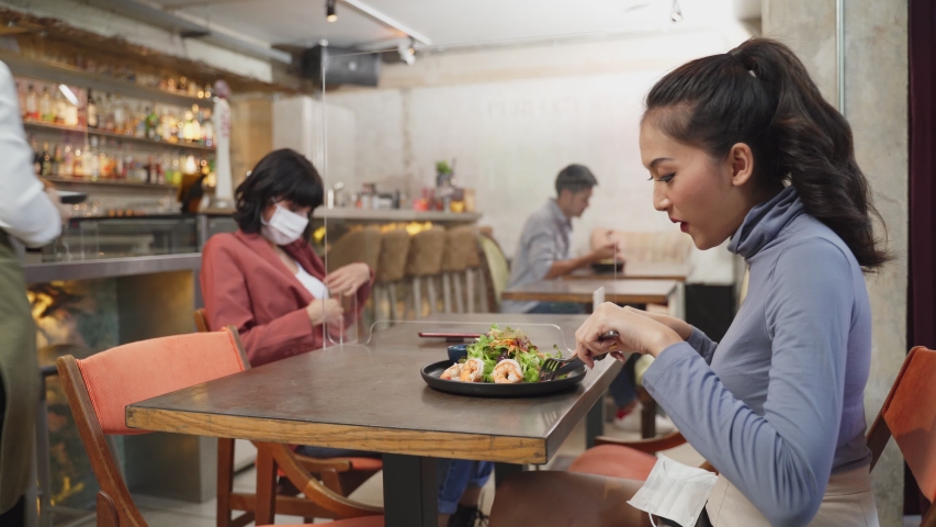 Asian customer woman dining in restaurant. Waitress with face mask serving food to customer sit on social distancing table for new normal lifestyle in restaurant after coronavirus covid-19 pandemic. Royalty-Free Stock Footage #1058264875