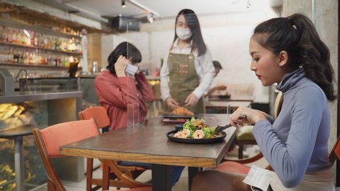 Asian customer woman dining in restaurant. Waitress with face mask serving food to customer sit on social distancing table for new normal lifestyle in restaurant after coronavirus covid-19 pandemic.