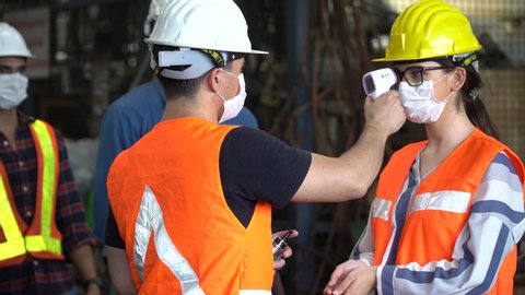 foreman wear protective face mask scanning body temperature measuring fever by thermometer and using  hand sanitizer alcohol gel to worker distancing before entering working in Industrial plant