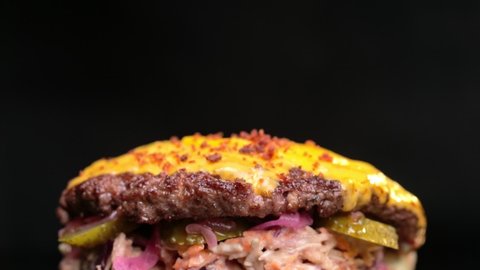 Making burger. Chef hands in black gloves are closing top bun of burger with oriental salad, cucumbers, onion and big tasty cutlet with melted cheese. Close up view. Camera zoom out. Black background.