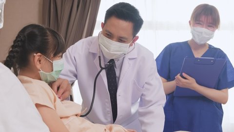 Asian doctor measure heart rate by stethoscope on little kid patient on bed in recovery room. Man and woman wearing mask having examination on girl during covid pandemic. Young nurse talking to child.