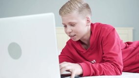 cute handsome and happy boy, teenager, sitting or lying on the bed in the room and watching videos laughing. learning lessons, schoolboy looking at laptop. the guy browses the internet and writes. 