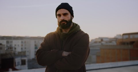 Portrait adult bearded man on rooftop at sunset wearing jacket and beanie hat looking confident in urban city background.Carefree holiday and happy active lifestyle.