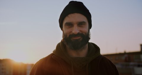 Portrait smiling adult bearded man on rooftop at sunset wearing jacket and beanie hat looking confident in urban city background.Carefree holiday and happy active lifestyle.