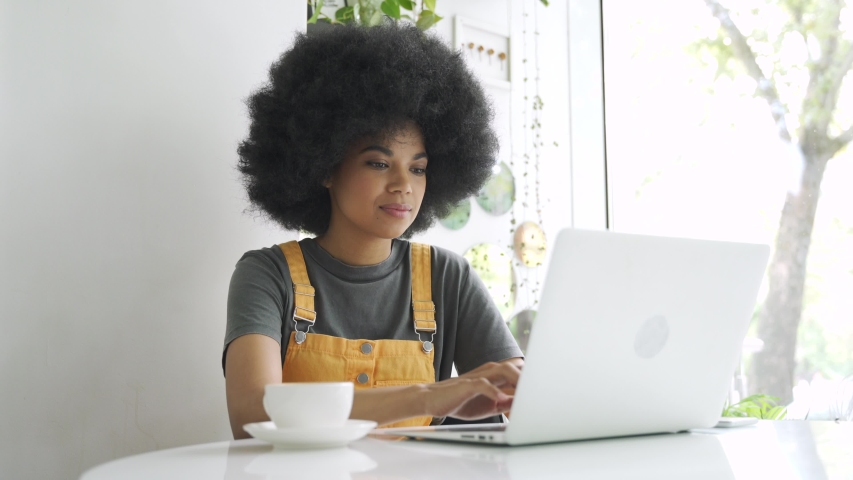 Happy stylish hipster gen z African American female student with afro hair using laptop computer, drinking coffee sitting at table at home, in cafe, remote studying, elearning or working in internet. Royalty-Free Stock Footage #1058278531