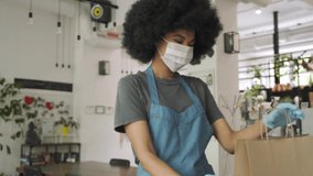 African American female cafe worker wears face mask and gloves giving takeaway food bag to customer. Mixed race waitress holding takeout order standing in coffee shop restaurant with take away client. - Powered by Shutterstock - Get 15% off with code: PIKWIZARD15
