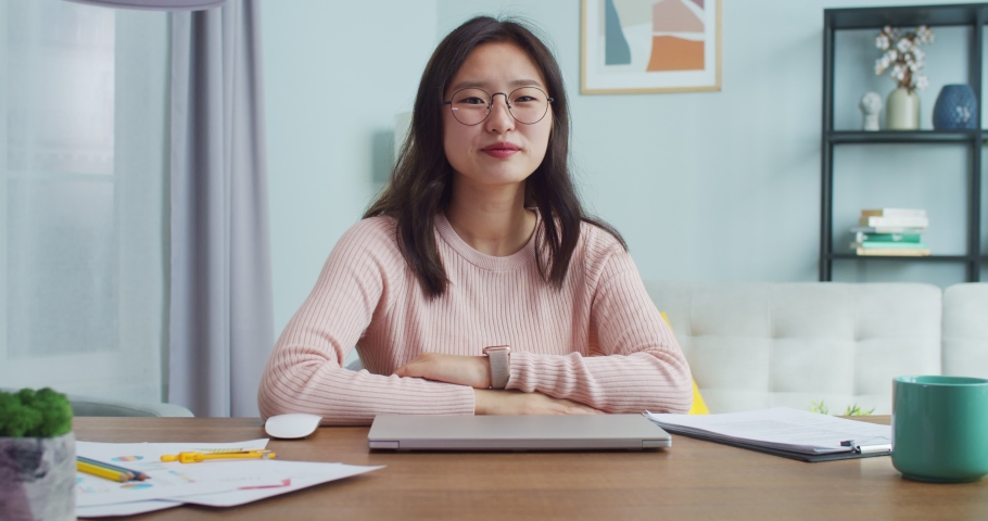 Pretty young Asian woman sitting at table in room with modern minimalist interior. Beautiful female student in glasses looking and smiling at camera. Distance learning and working. | Shutterstock HD Video #1058280751