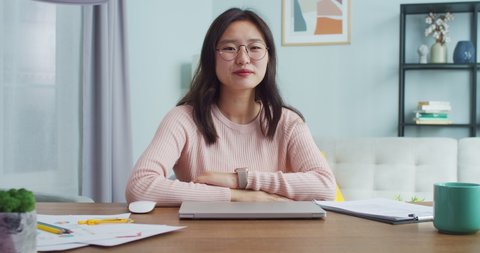 Pretty young Asian woman sitting at table in room with modern minimalist interior. Beautiful female student in glasses looking and smiling at camera. Distance learning and working.