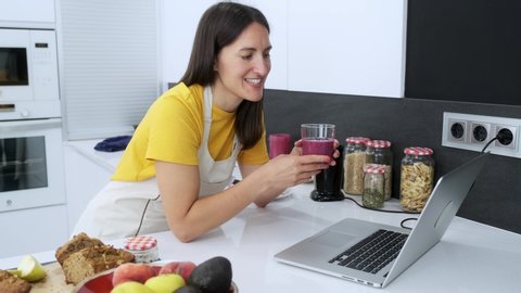 Video of beautiful young nutritionist woman having an online video call via laptop computer with a friend to showing detox handmade smoothie in the kitchen at home.
