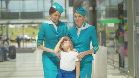 Charming little girl standing with stewardesses next to airport and looking at plane takeoff. Portrait of happy Caucasian child enjoying sunny day with professional flight attendants before departure.