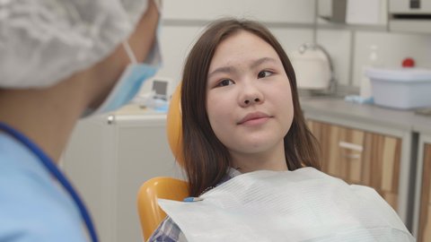 Rear view of Asian girl lying on dental chair in clinic and telling female dentist sitting in front of her about her teeth problems
