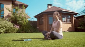 Online meditation lesson. Young woman in sportswear walking in lotus position looking at laptop screen and giving online yoga lessons while meditating outdoors in the morning
