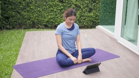 Asian woman stretching body and yoga on deck outside home and watching live or video tutorial online via tablet computer. Activity during quarantine and social distance new normal concept.