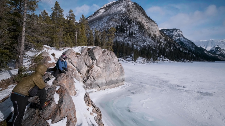 Young Hiking Couple Enjoys a Breathtaking Winter Lookout in Snowy Mountains of Banff National Park at Lake Minnewanka Alberta Canada. Sunny Day Pan Right Handheld, Two White People in Down Parka Coat