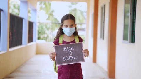 Young girl in a medical mask coming front by looking camera and showing by holding back to school signage board at corridor concept of school reopen. lifestyle and new normal.back to school concept.