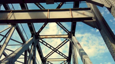 iron construction of bridge against blue sky with white clouds. driving under bridge, view of bridge from below. Silhouette of crossing steel beams. Bridge details against bright blue sky.