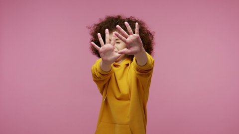 I'm afraid! Frustrated scared young woman afro hairstyle in hoodie stretching out hands, stop gesture to hide from fear, looking terrified afraid, panic attack. indoor  isolated on pink background
