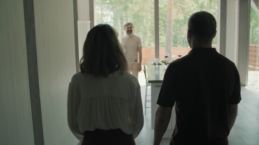 Tracking of happy young couple walking into room of modern house with panoramic windows and hugging senior people, then sitting down at dinner table to eat lunch