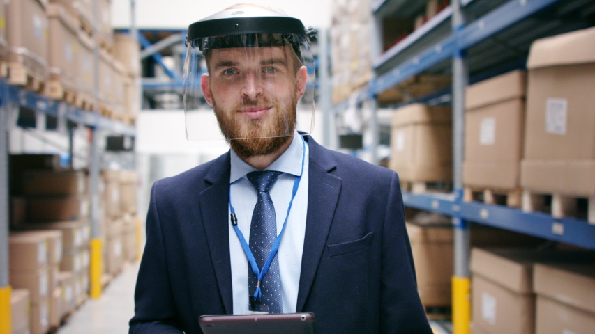 Man manager with protective face shield and tablet walking in warehouse.