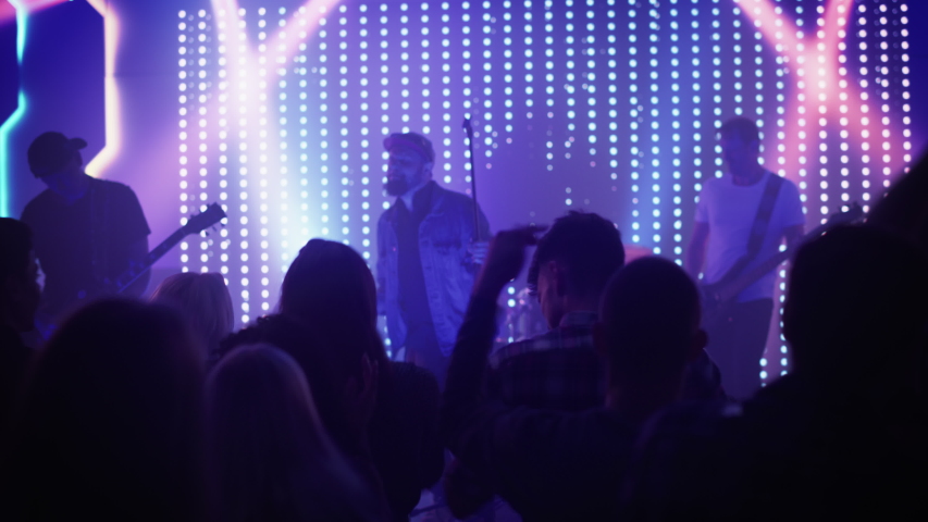 Rock Band with Guitarists and Drummer Performing at a Concert in a Night Club. Front Row Crowd is Partying. Silhouettes of Fans Raise Hands in Front of Bright Colorful Strobing Lights on Stage. Royalty-Free Stock Footage #1058308843