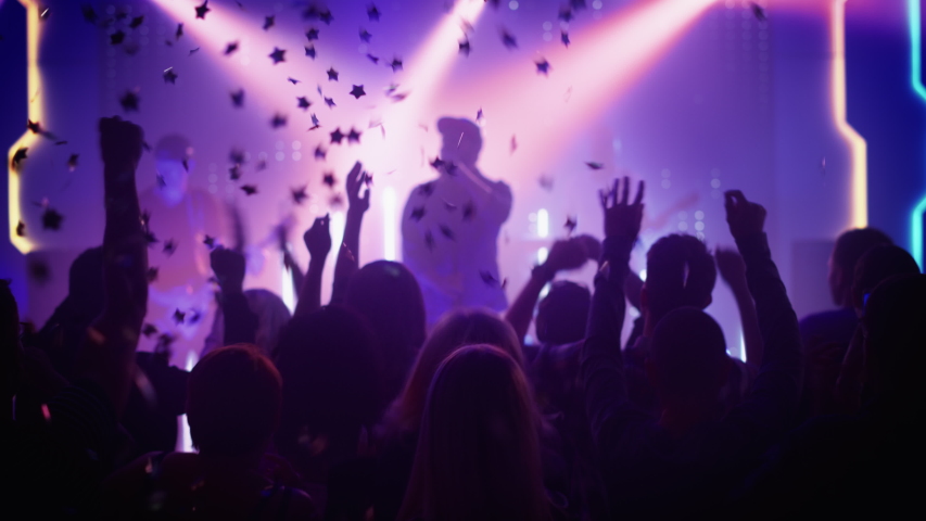Rock Band with Guitarists and Drummer Performing at a Concert in a Night Club. Front Row Crowd is Partying. Silhouettes of Fans Raise Hands in Front of Bright Colorful Strobing Lights on Stage. Royalty-Free Stock Footage #1058308858