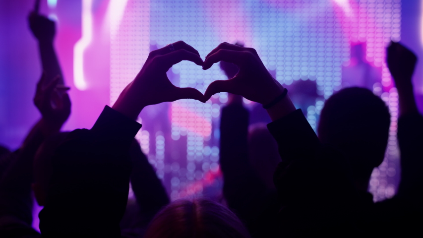 Person is Making a Heart Sign Gesture and Holding Hands Up at a Performance. Rock Band Playing a Song at a Concert in a Night Club on Stage with Bright Colorful Strobing Lights. Royalty-Free Stock Footage #1058308903