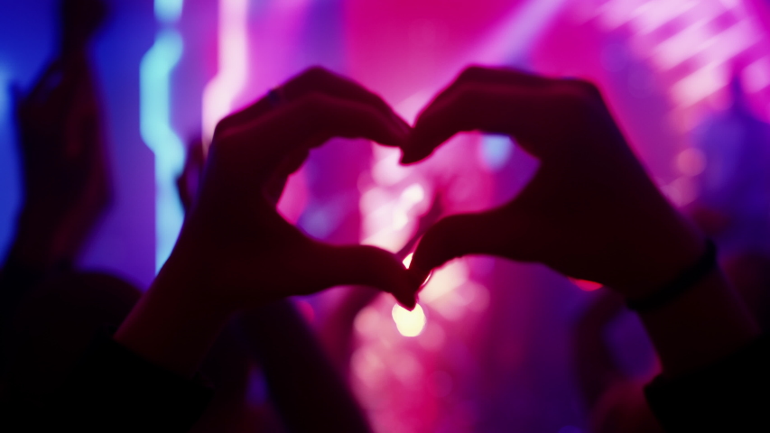 Person is Making a Heart Sign Gesture and Holding Hands Up at a Performance. Rock Band Playing a Song at a Concert in a Night Club on Stage with Bright Colorful Strobing Lights. Royalty-Free Stock Footage #1058308921
