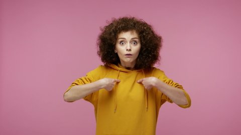 Wow, this is me! Amazed excited young woman afro hairstyle in hoodie pointing herself, looking with surprised astonished expression, shocked by own success. indoor  isolated on pink background