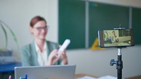 young woman teaches lesson in an online school, teacher in glasses with cards in hands uses video camera on mobile phone for recording sitting at table