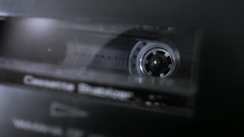An audio cassette spools tape in extreme macro. Shallow depth of field with focus on the rorating spool. Suggests an interrogation or phonetap. 
