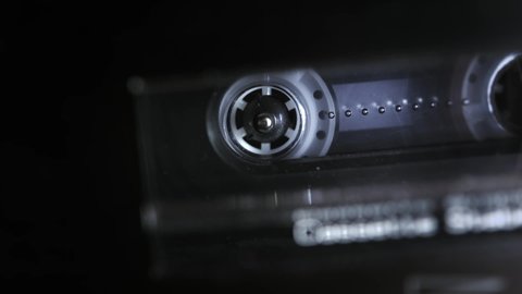 An audio cassette spools tape in extreme macro. Shallow depth of field with focus on the rorating spool. Suggests an interrogation or phonetap. 