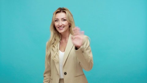 Friendly positive middle aged blond woman in stylish suit waving hand looking at camera with toothy smile, saying hi, greeting. Indoor studio shot isolated on blue background
