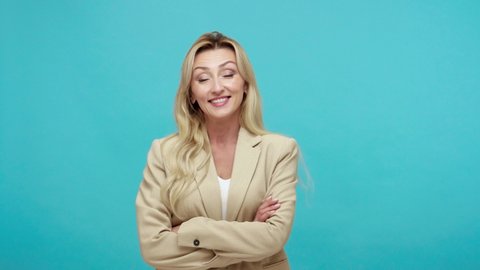 Positive self-confident blonde woman in business style jacket crossing her arms on chest and smiling, showing her arrogance. Indoor studio shot isolated on blue background