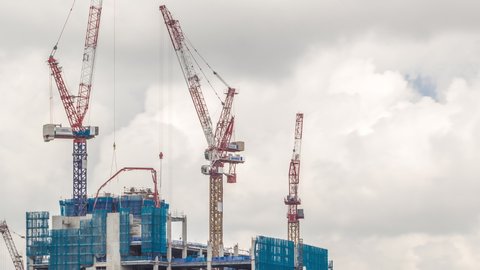Working cranes on a modern office building under construction against cloudy sky in Singapore timelapse. Asian urban development and construction concept. Industrial background.