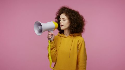 Funny young woman afro hairstyle in hoodie talking with megaphone, proclaiming news, loudly announcing advertisement, warning using loudspeaker to shout speech. studio shot isolated on pink background