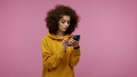 young woman afro hairstyle in hoodie scrolling social network using mobile phone and looking at camera with surprised expression, shocked by device or app. indoor   isolated on pink background