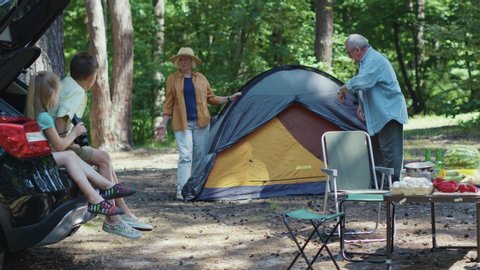 Grandparents succeed to put up camping tent in the forest. They calling their adoorable grandchildren running quickly. Caucasian family. Recreation. Outdoor activities.